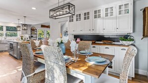 Una cocina moderna con armarios blancos, una mesa de comedor de madera, sillas de mimbre y una lámpara colgante.