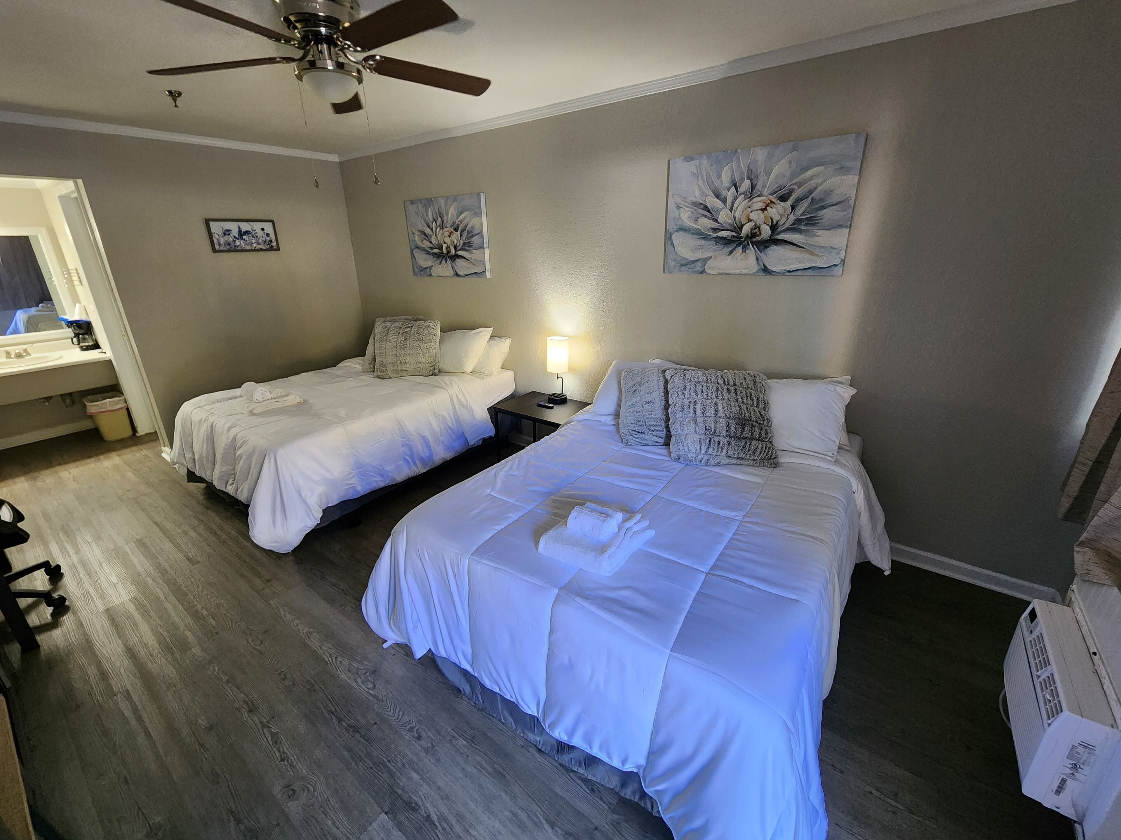 A hotel room with two beds, a ceiling fan, artwork on the walls, a bedside table with a lamp, and a window with a view.