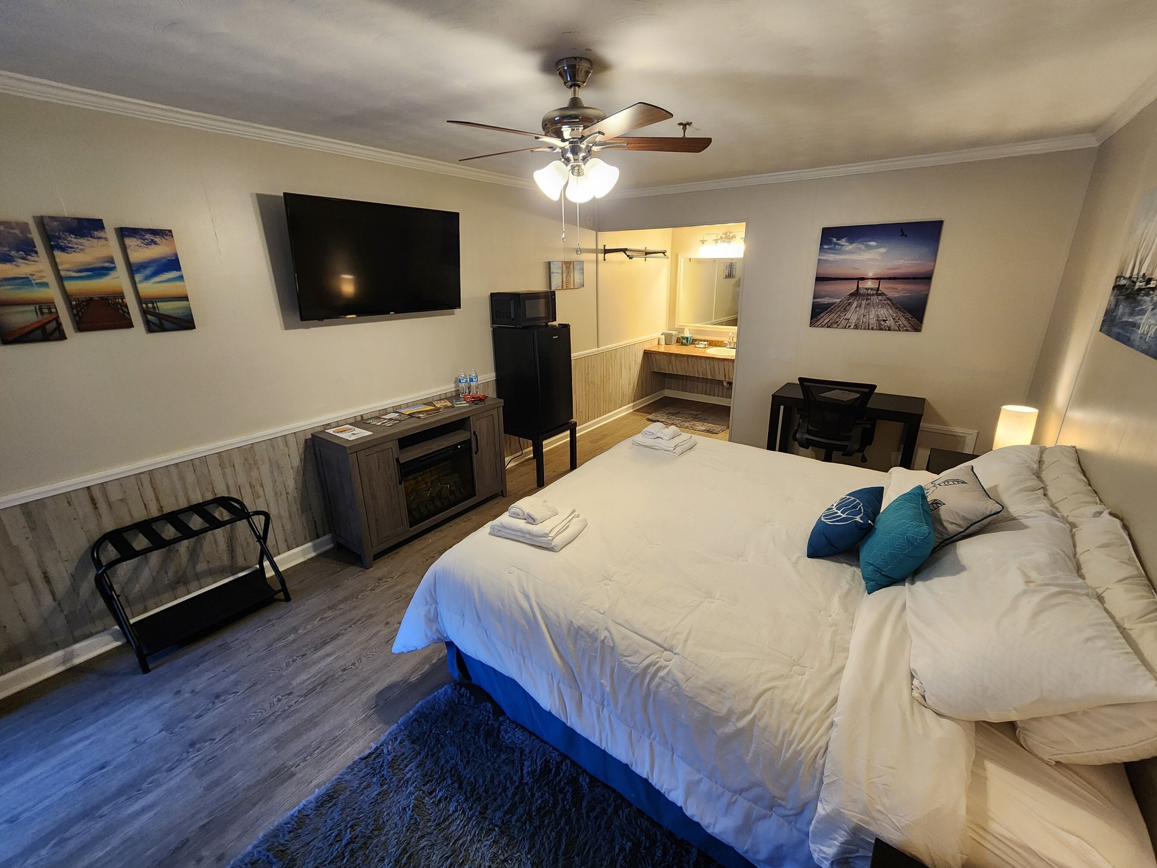 A hotel room with a bed, a flat-screen TV, a ceiling fan, a fireplace, a small table, and artwork on the walls.