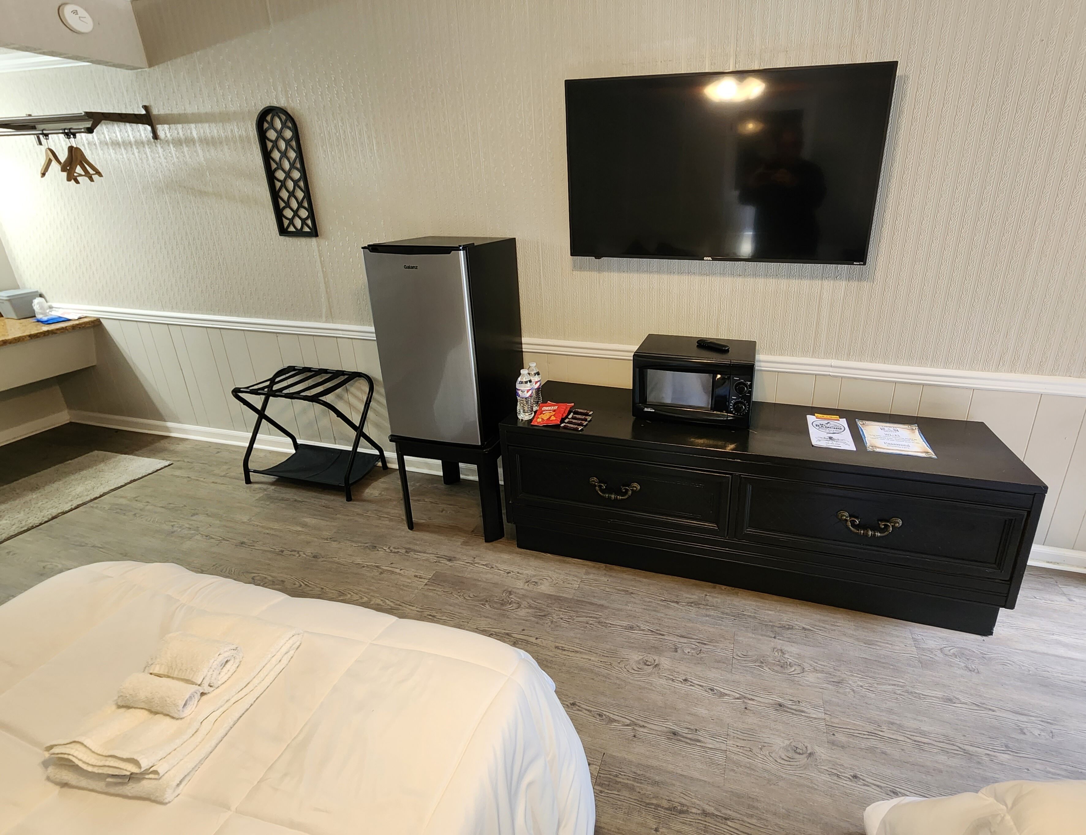 A hotel room with a bed, a flat-screen TV mounted on the wall, a minibar, a small table with a microwave, and a black storage unit with drawers.