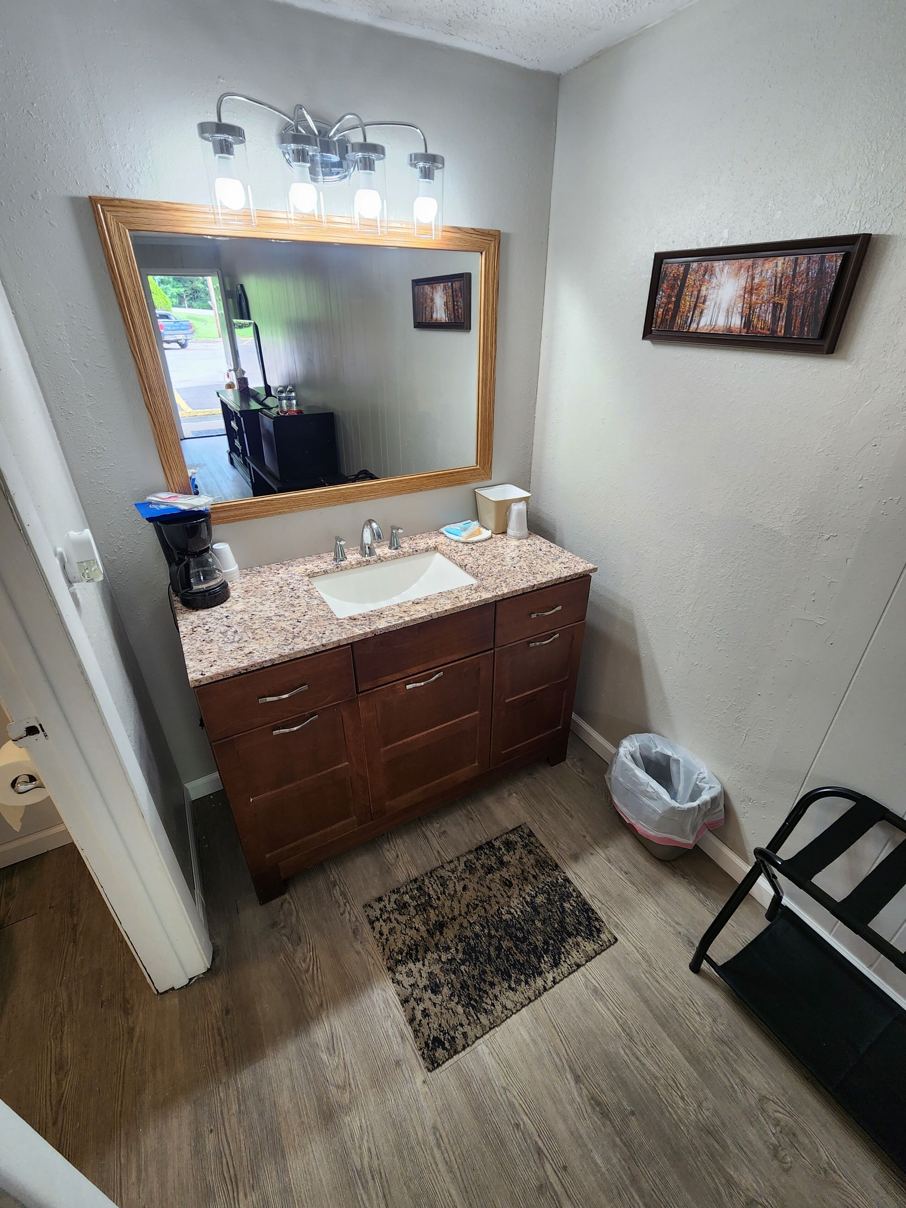 A bathroom with a granite countertop sink, wooden vanity, a large mirror with lights, a framed picture on the wall, and a small rug.