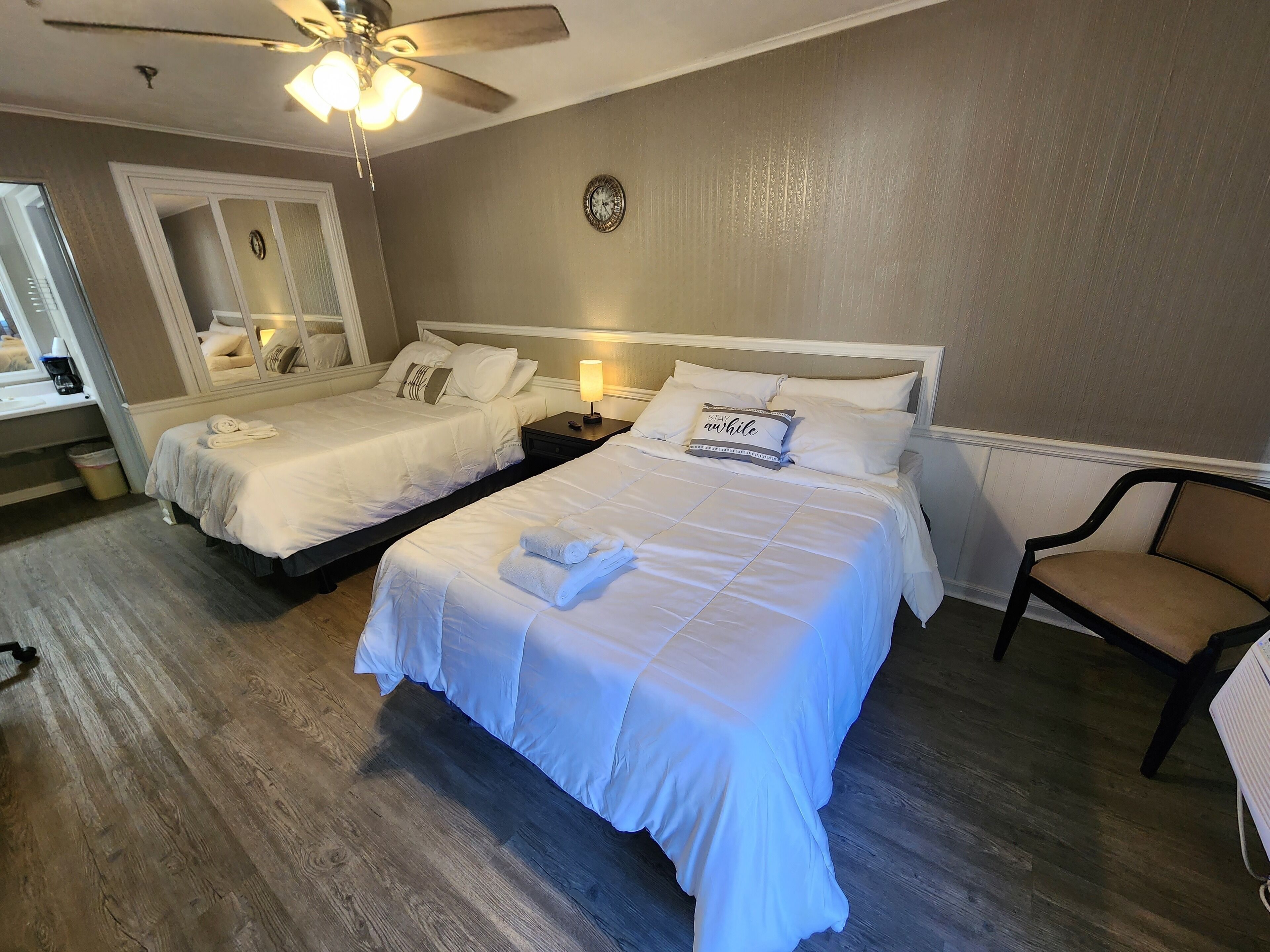 A hotel room with two beds, a ceiling fan, a clock, and a chair.