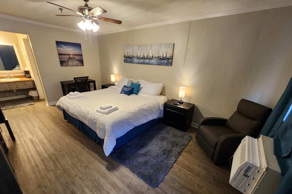 Een keurig opgemaakt slaapkamer met een groot bed, nachtkastjes, een stoel en een plafondventilator.