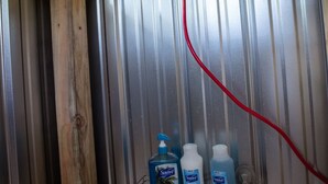 A shower area with a wooden ceiling and metal walls, equipped with a handheld showerhead and a metal shelf holding cleaning supplies. - Cozy campsite with creek access! (Landisburg)