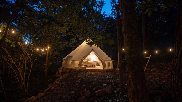 A tent with a bed and a table, surrounded by trees and rocks, with string lights. - Cozy campsite with creek access! (Landisburg)