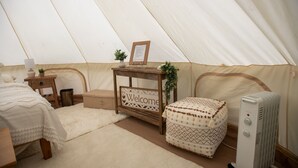 A tented room with a bed, a wooden nightstand, a welcome sign, a small table with a plant, a patterned ottoman, and a heater. - Cozy campsite with creek access! (Landisburg)