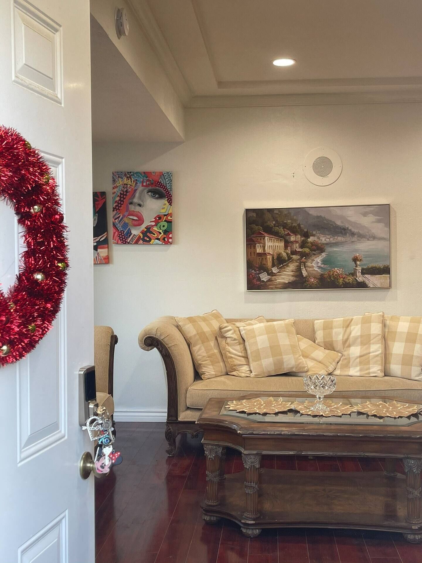A living room with a beige sofa, a glass coffee table, and a red tinsel wreath on the door.
