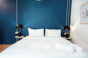 A neatly made bed with white linens and pillows, two bedside tables with black lamps, and a framed artwork on the wall. - Selina Kinneret (Migdal)