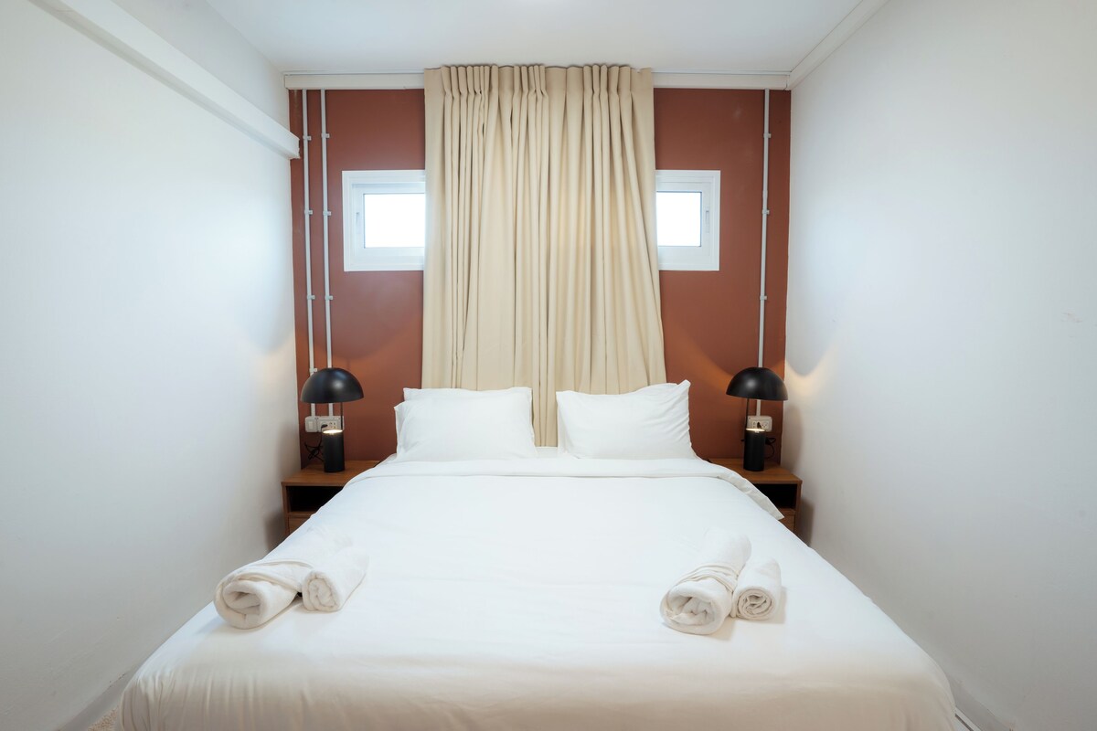 A neatly made bed with white linens and two rolled towels, a window with beige curtains, and two bedside lamps on wooden nightstands.