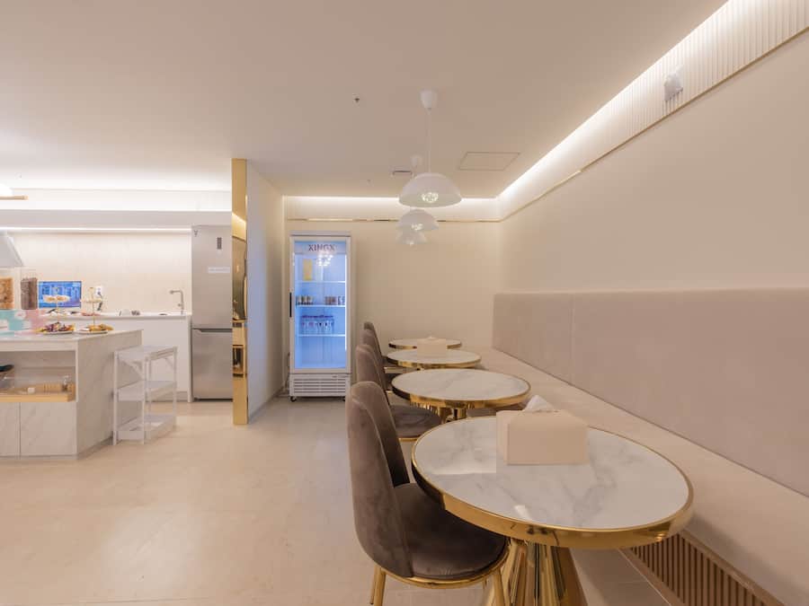 Un café moderno con mesas de mármol y sillas acolchadas, una barra con vitrina expositora y un refrigerador al fondo.