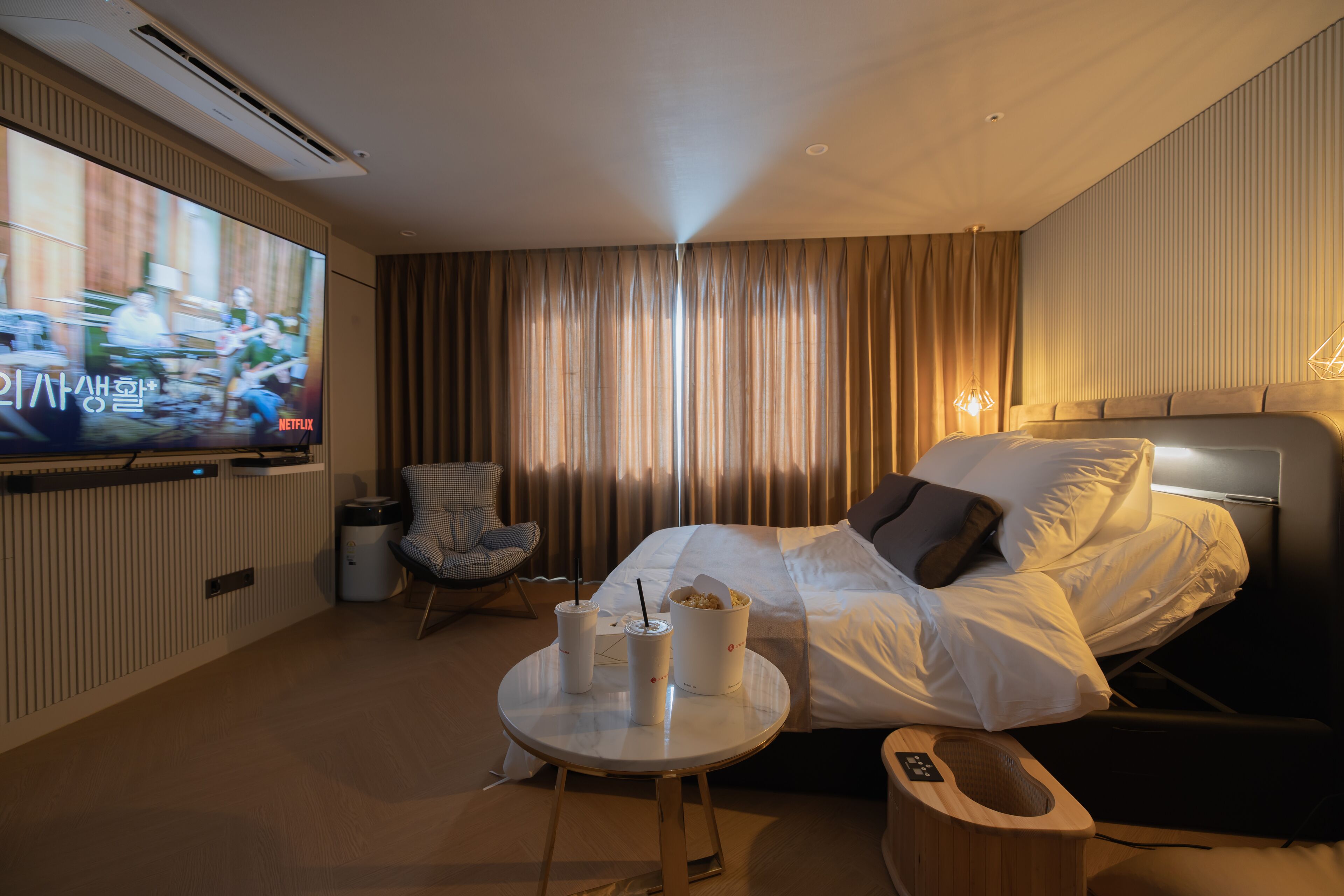 A hotel room with a bed, a TV, a small table with drinks, a chair, and a bedside table with a remote control.