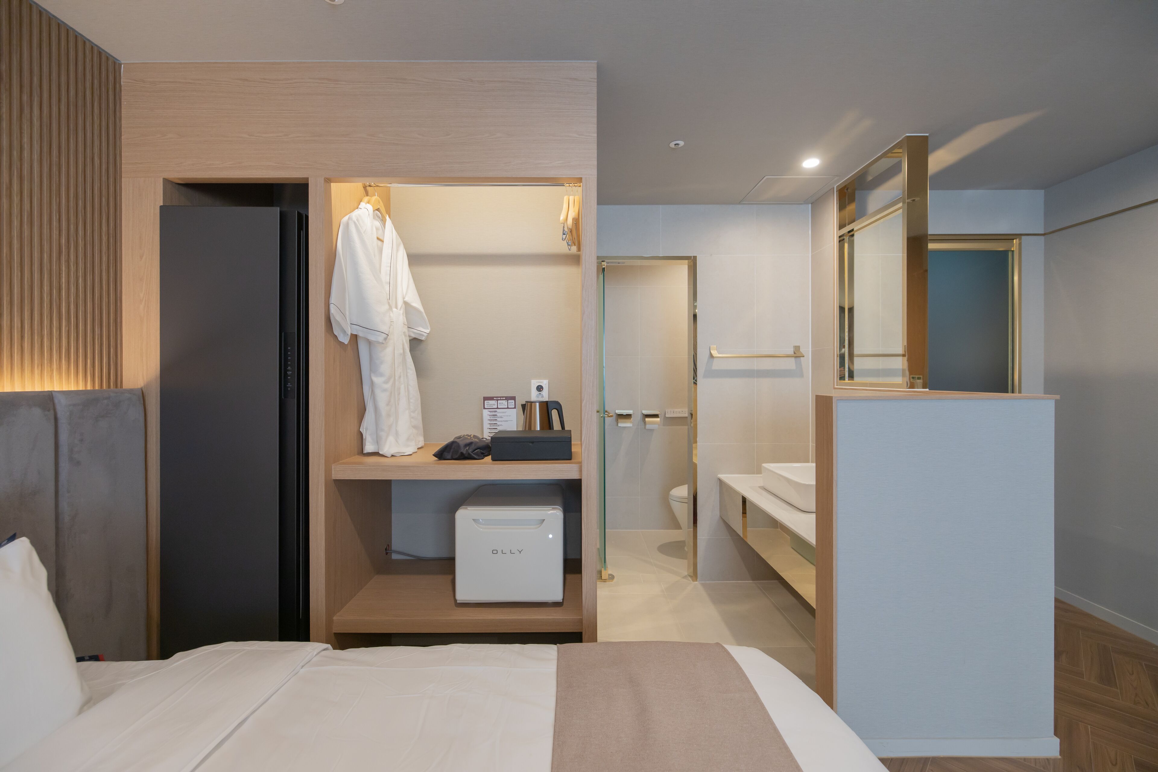 A modern hotel room with a bed, a built-in wardrobe, a small kitchenette, and a bathroom with a toilet and sink.