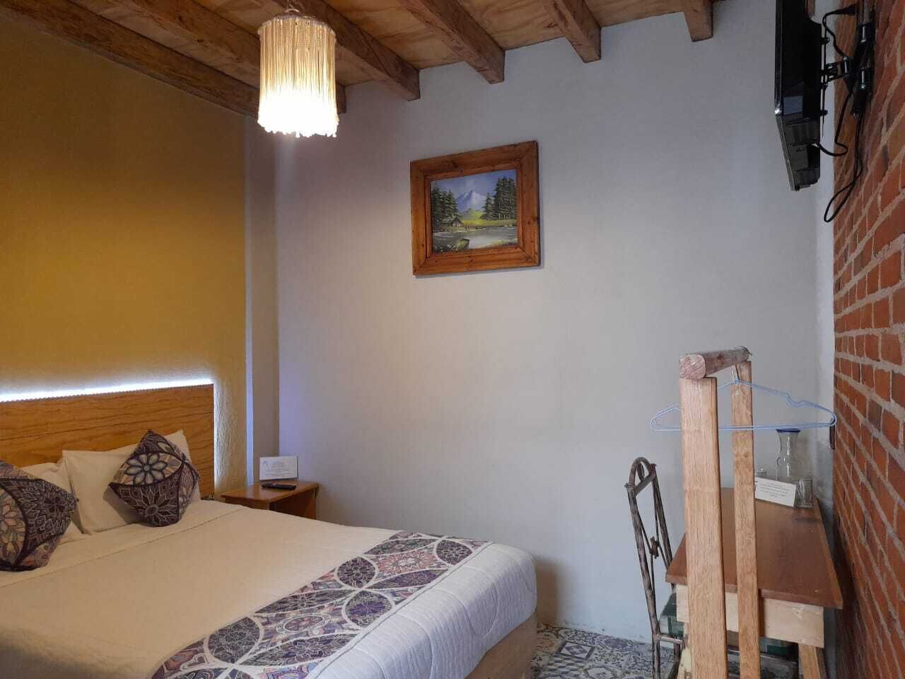 A bedroom with a wooden headboard, a bed with patterned pillows, a hanging light fixture, a painting on the wall, and a wooden chair.