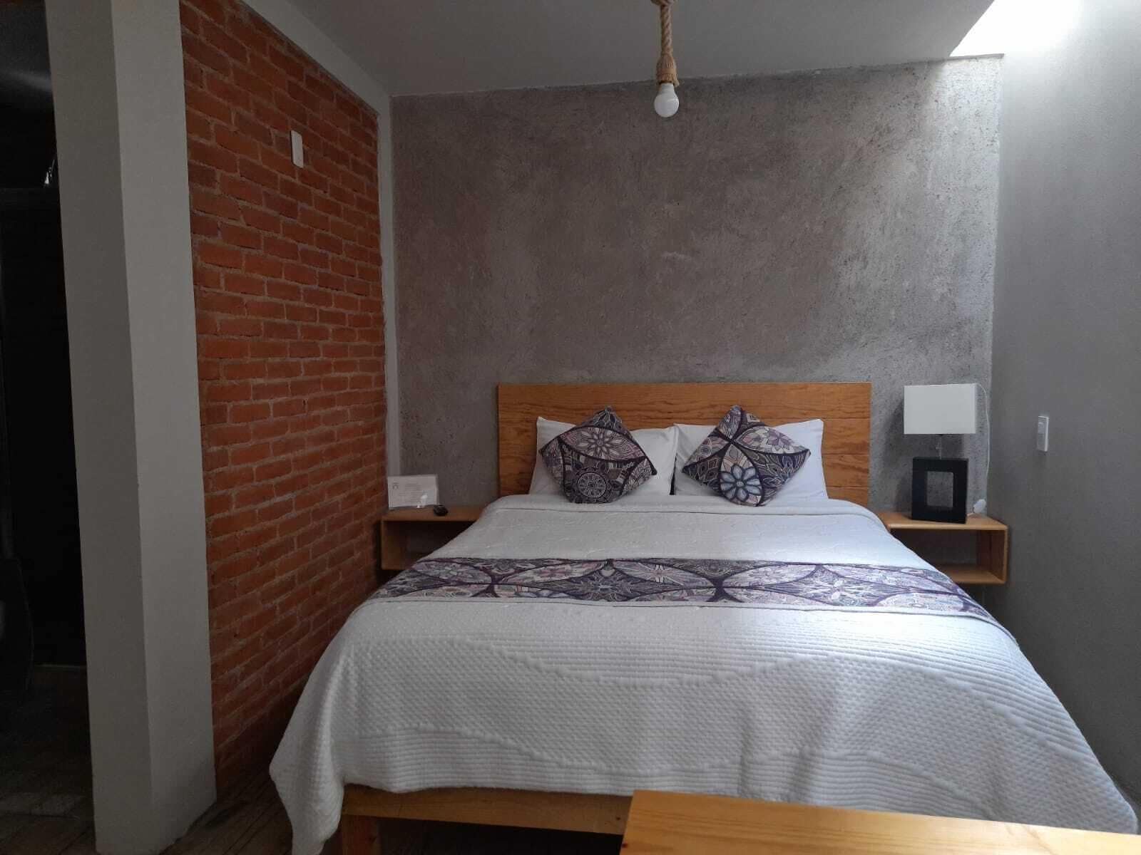 A bedroom with a brick wall, a wooden headboard, a patterned bedspread, two pillows, a bedside table with a lamp, and a hanging light fixture.