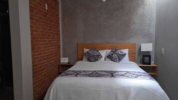 A bedroom with a brick wall, a wooden headboard, a patterned bedspread, two pillows, a bedside table with a lamp, and a hanging light fixture.