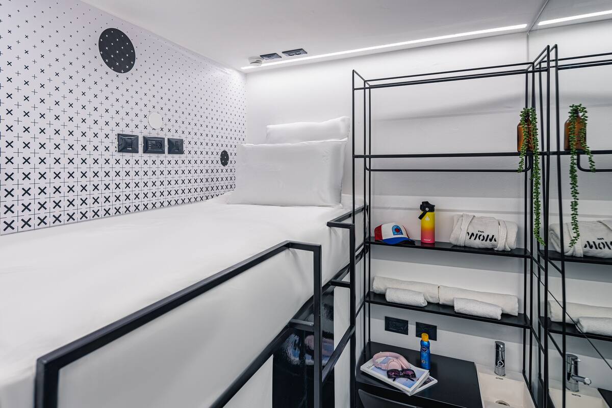 Ein modernes Hotelzimmer mit Wand in Weiß und Schwarz, einem schwarzen Metallregal mit Handtüchern und Pflegeprodukten, einem kleinen Schreibtisch mit einem Laptop und einem schwarzen Stuhl.