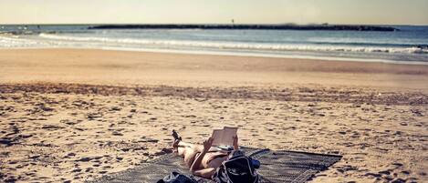 Una persona recostada sobre una toalla de playa con un libro, rodeada de equipo de playa.