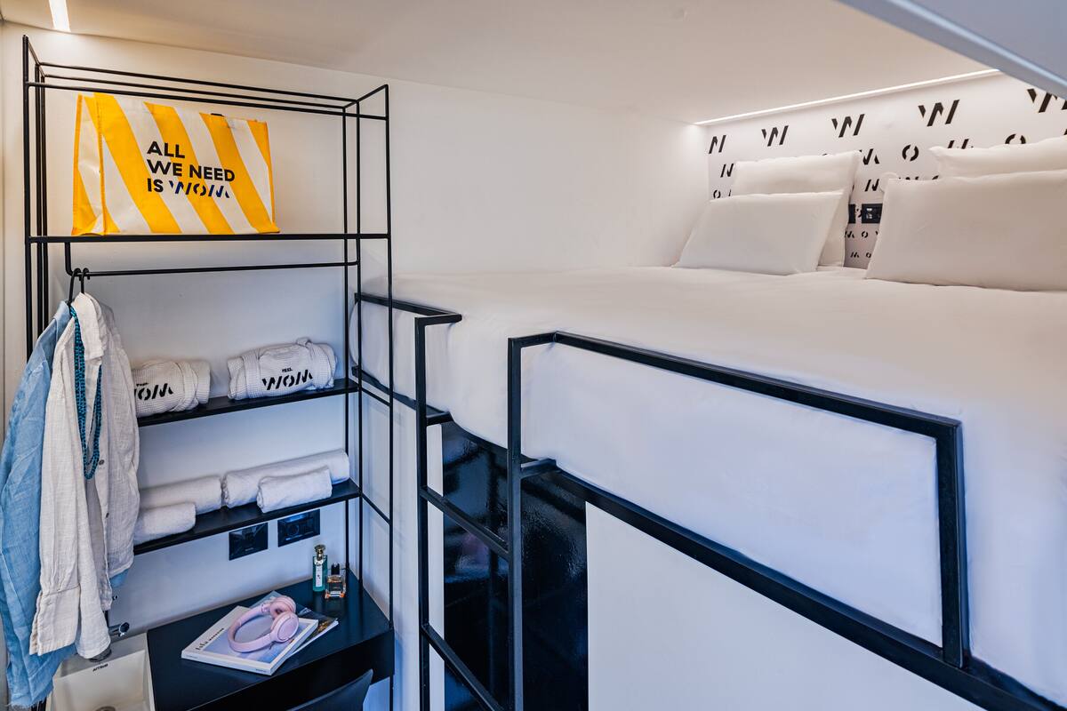 Ein kleines Hotelzimmer mit Etagenbett, Schreibtisch und Stuhl, Ablagefläche mit Handtüchern und einer Wand mit dekorativem Muster.