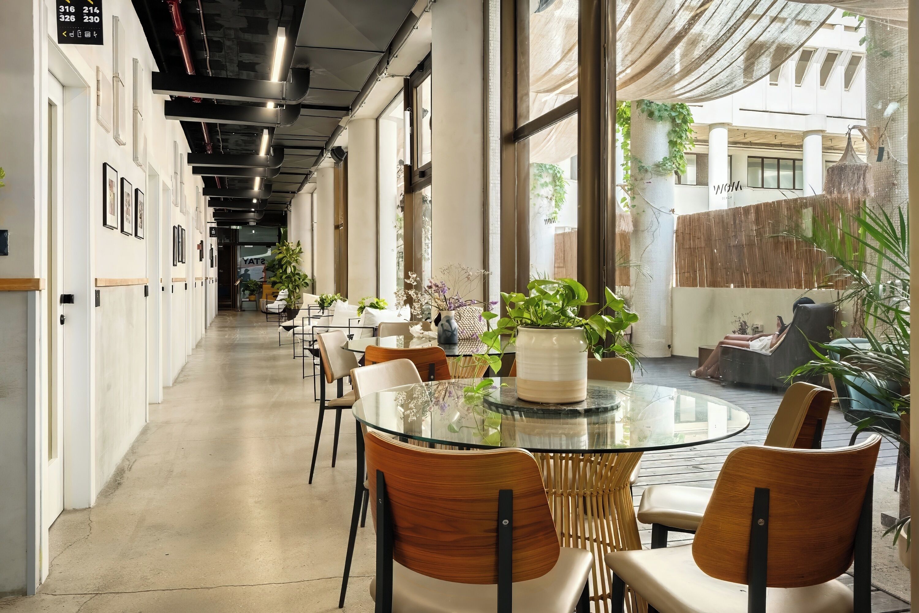 A modern indoor dining area with round glass tables and wooden chairs, large windows, and indoor plants.