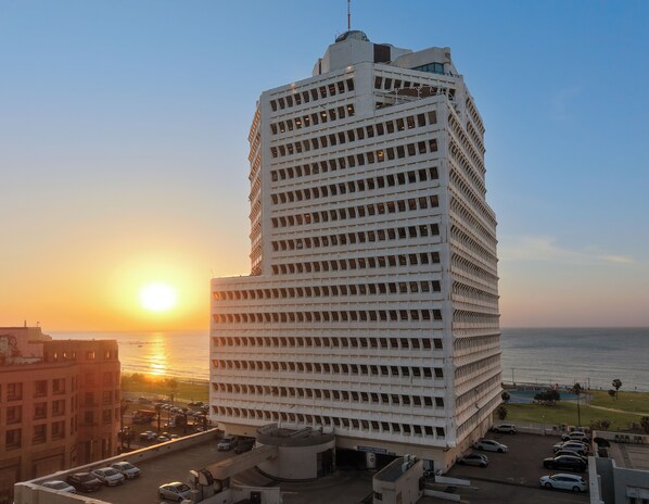 A high-rise building with a grid-like facade, a parking area in front, and a view of the ocean at sunset. - WOM Beach Pod Hotel  (Tel Aviv)