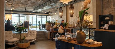 A modern cafe with a high ceiling, exposed beams, and large windows offering a view. There are seating areas with chairs and tables, and a counter with a barista.