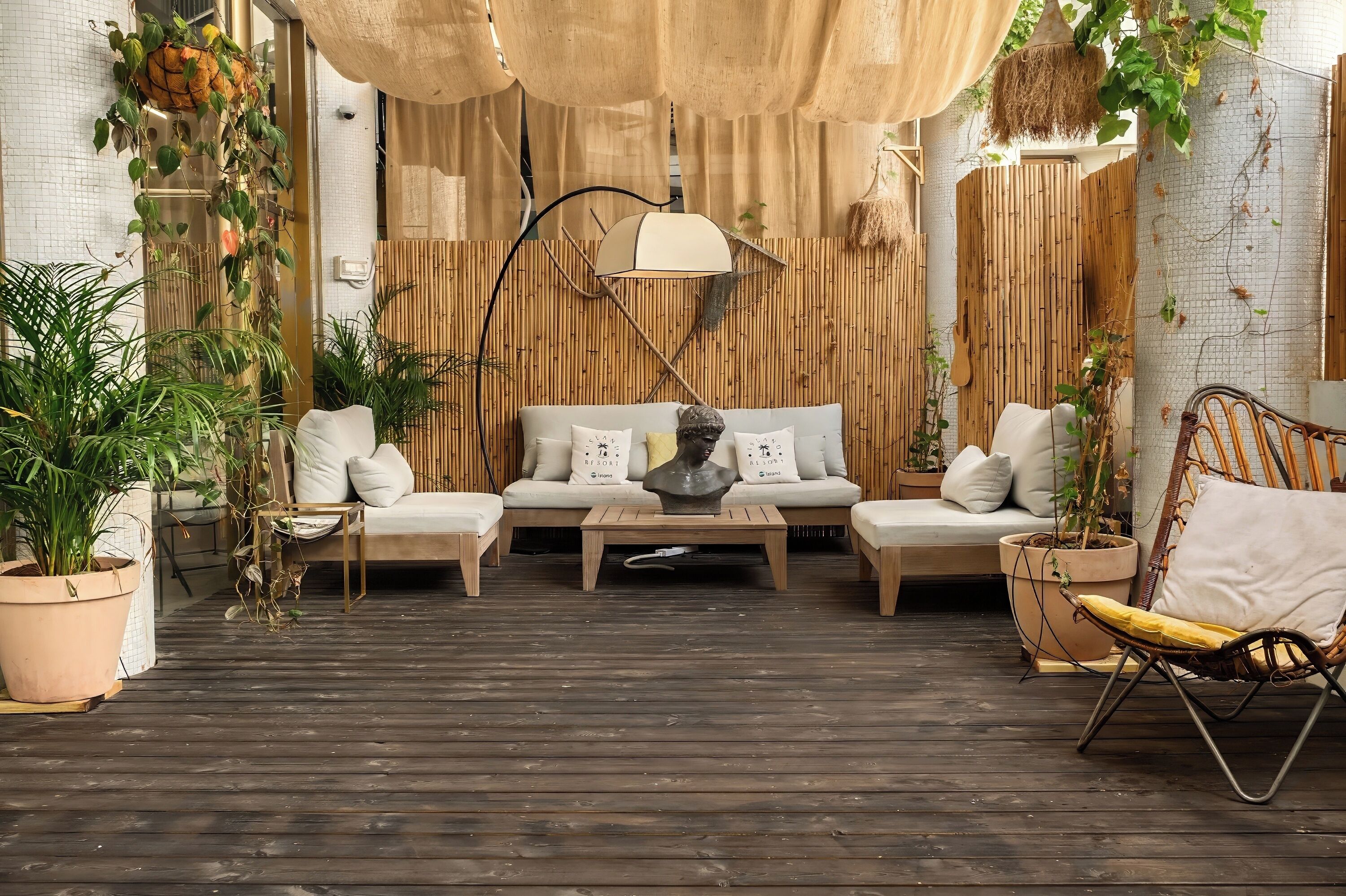 Uma área de estar ao ar livre acolhedora, com um sofá, cadeiras e uma mesa de centro, cercada por plantas em vasos e um painel de bambu.