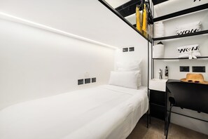 A minimalist hotel room with a single bed, a small desk, and a black chair. There are shelves with folded towels and toiletries. - WOM Beach Pod Hotel  (Tel Aviv)