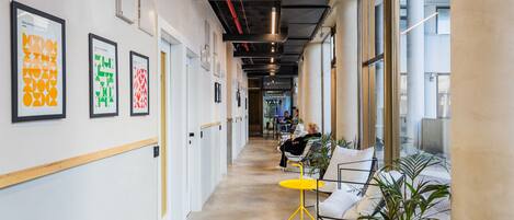 A modern office corridor with framed artwork on the walls, a yellow table, and potted plants.