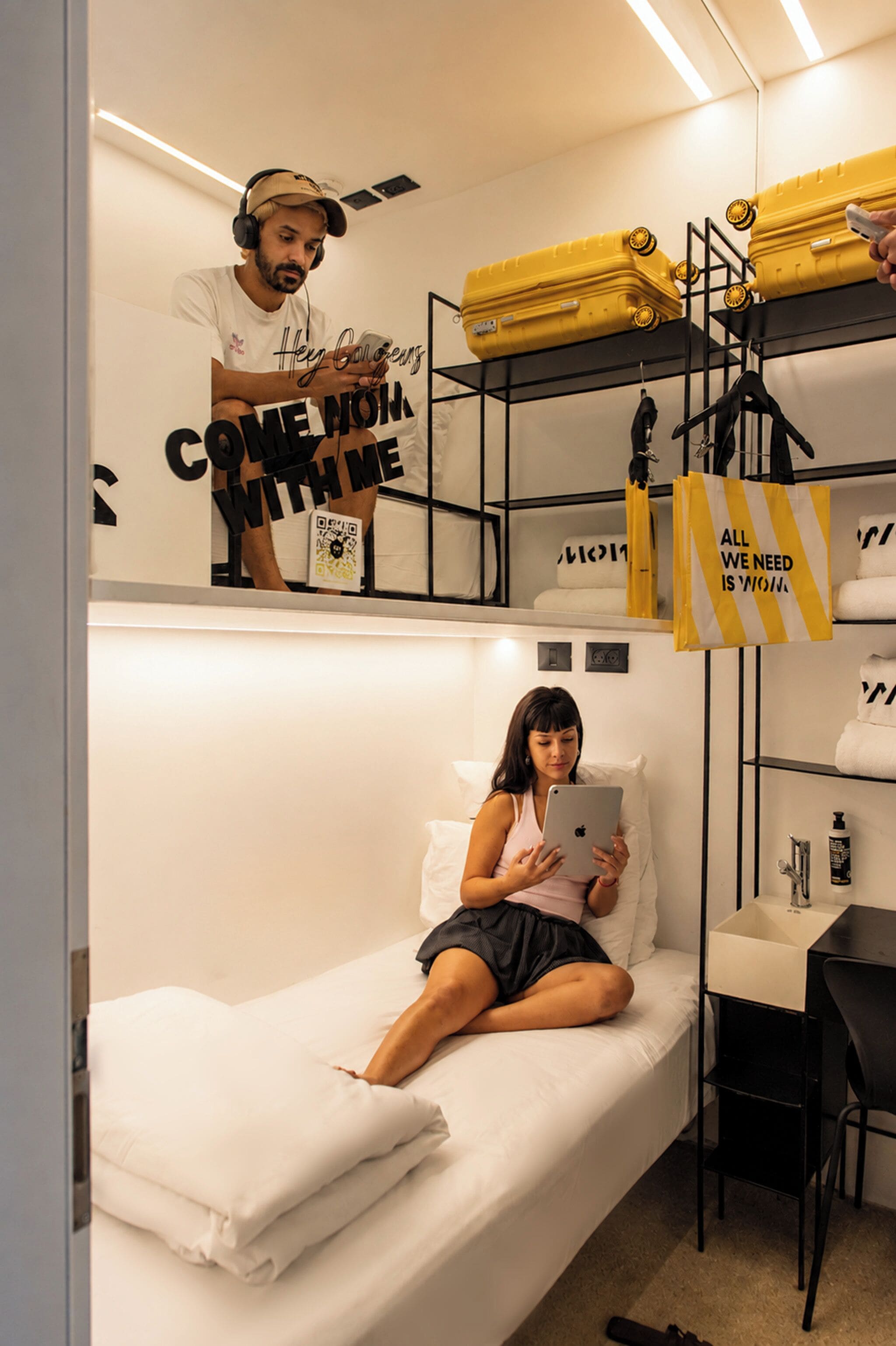 A bunk bed with a person sitting on the lower bunk, a suitcase on the upper bunk, and a small table with a chair.