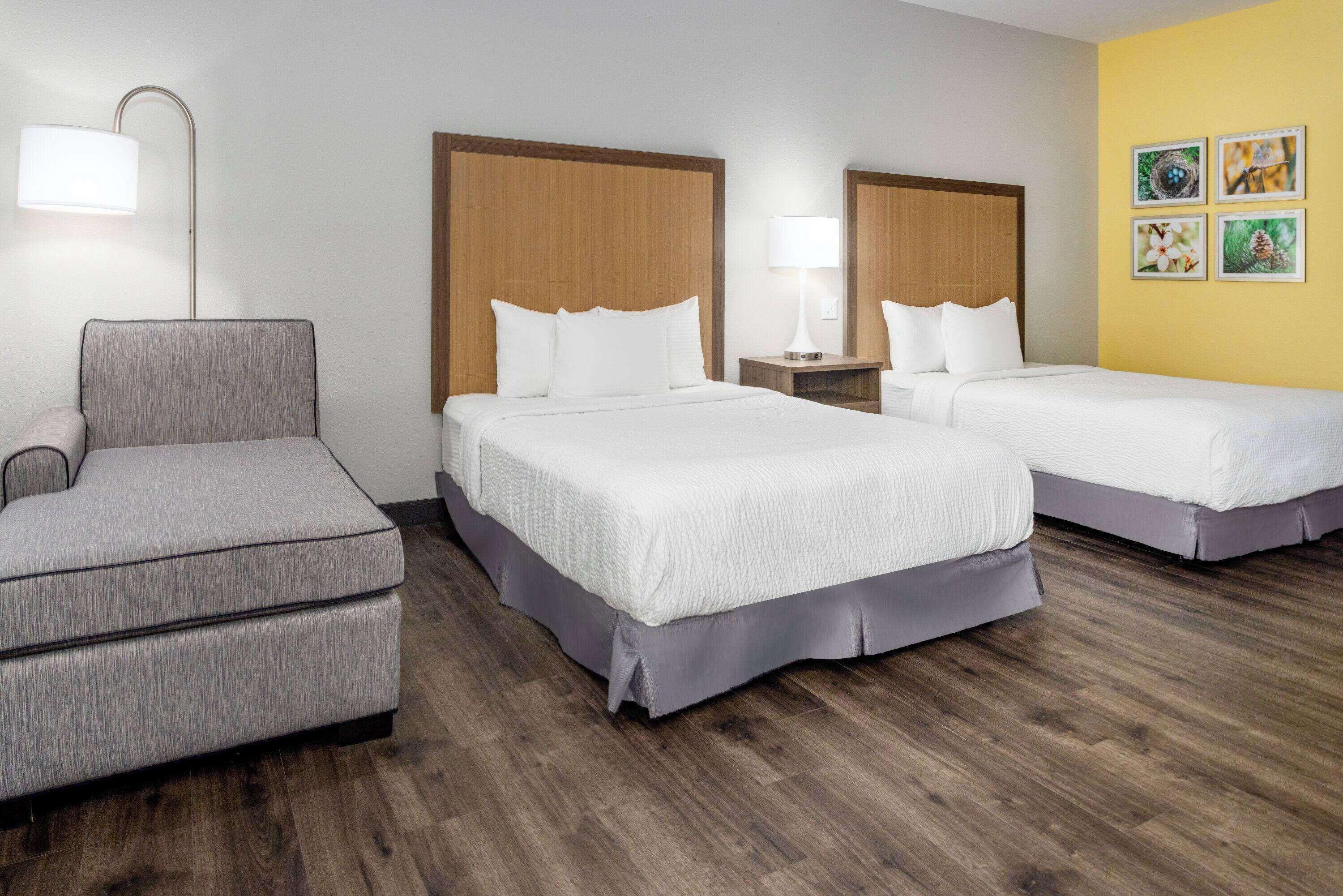 Habitación de hotel con dos camas, una silla, una mesita de noche y una lámpara.