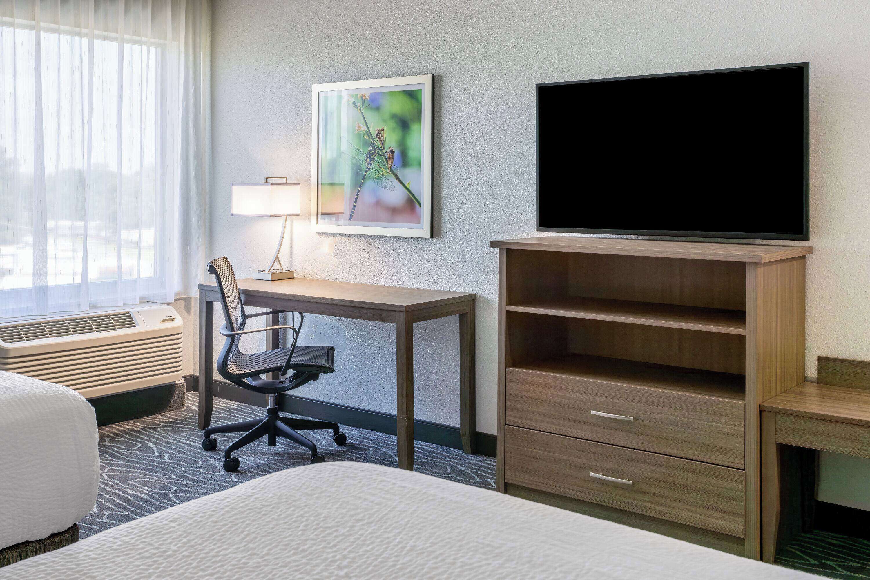 A hotel room with a bed, a desk with a chair, a television, and a wall-mounted shelf.