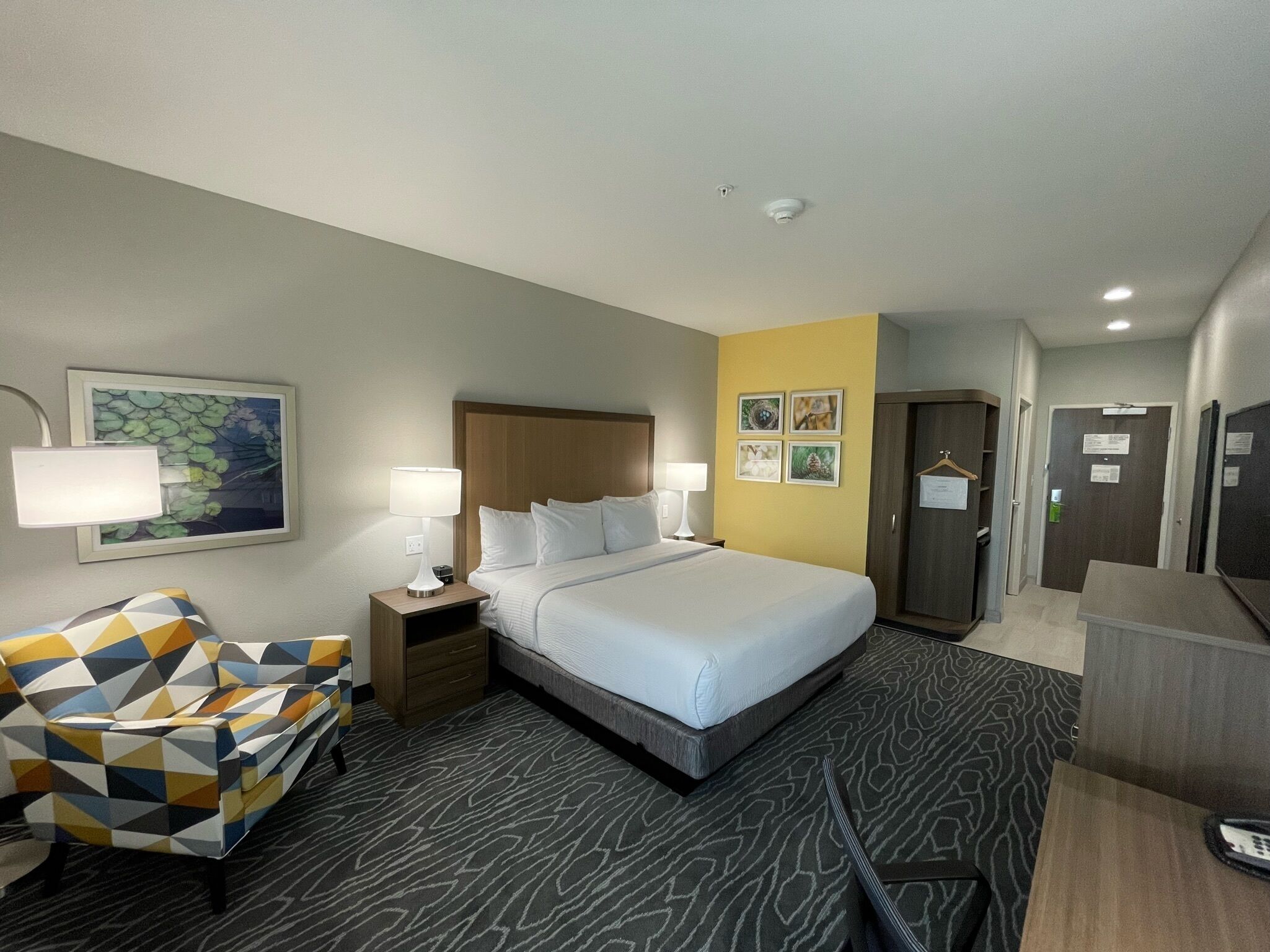 A hotel room with a large bed, two bedside tables with lamps, a chair, a wardrobe, and a door with a sign.