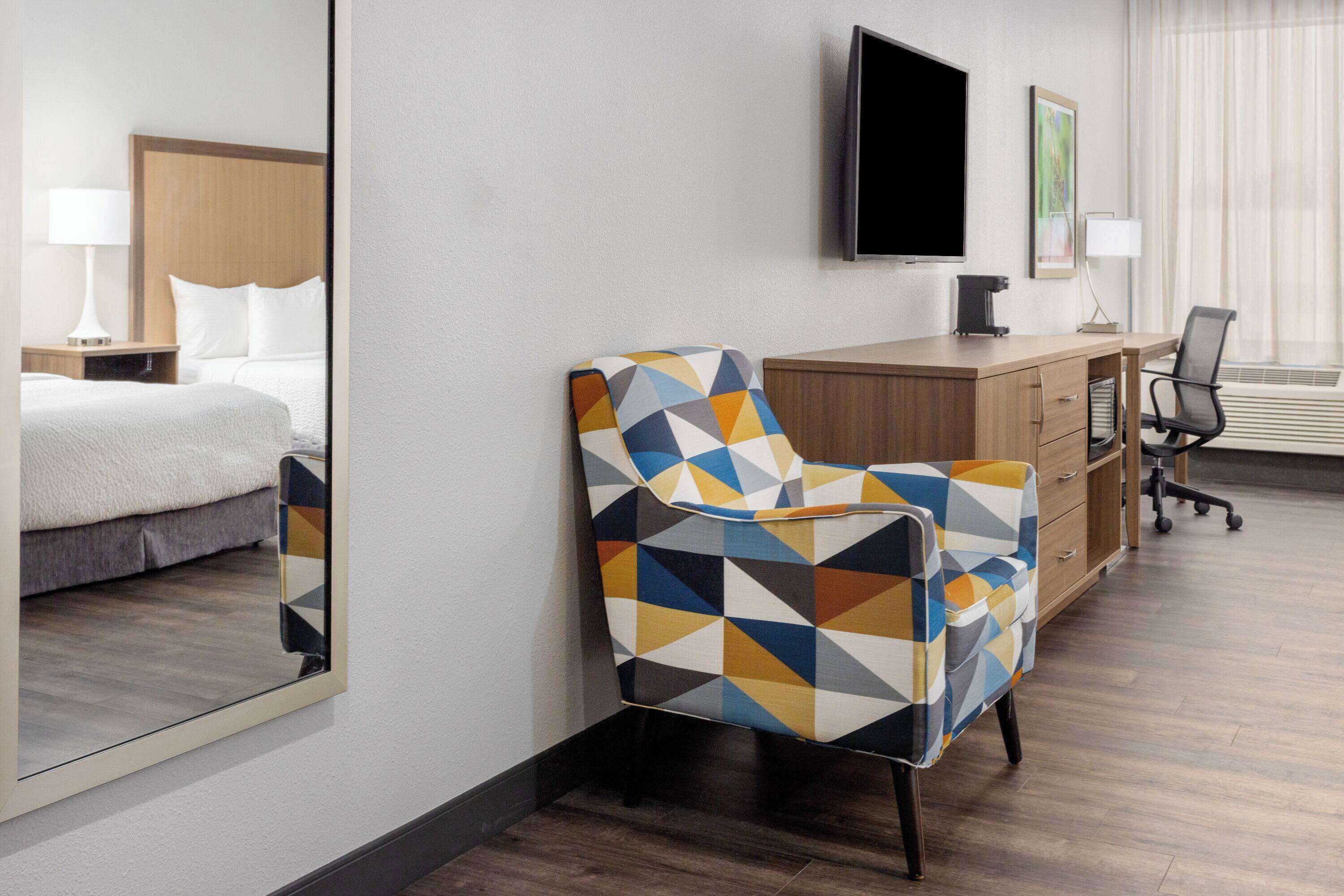 A hotel room with a bed, a desk, a chair, a TV, and a large mirror.