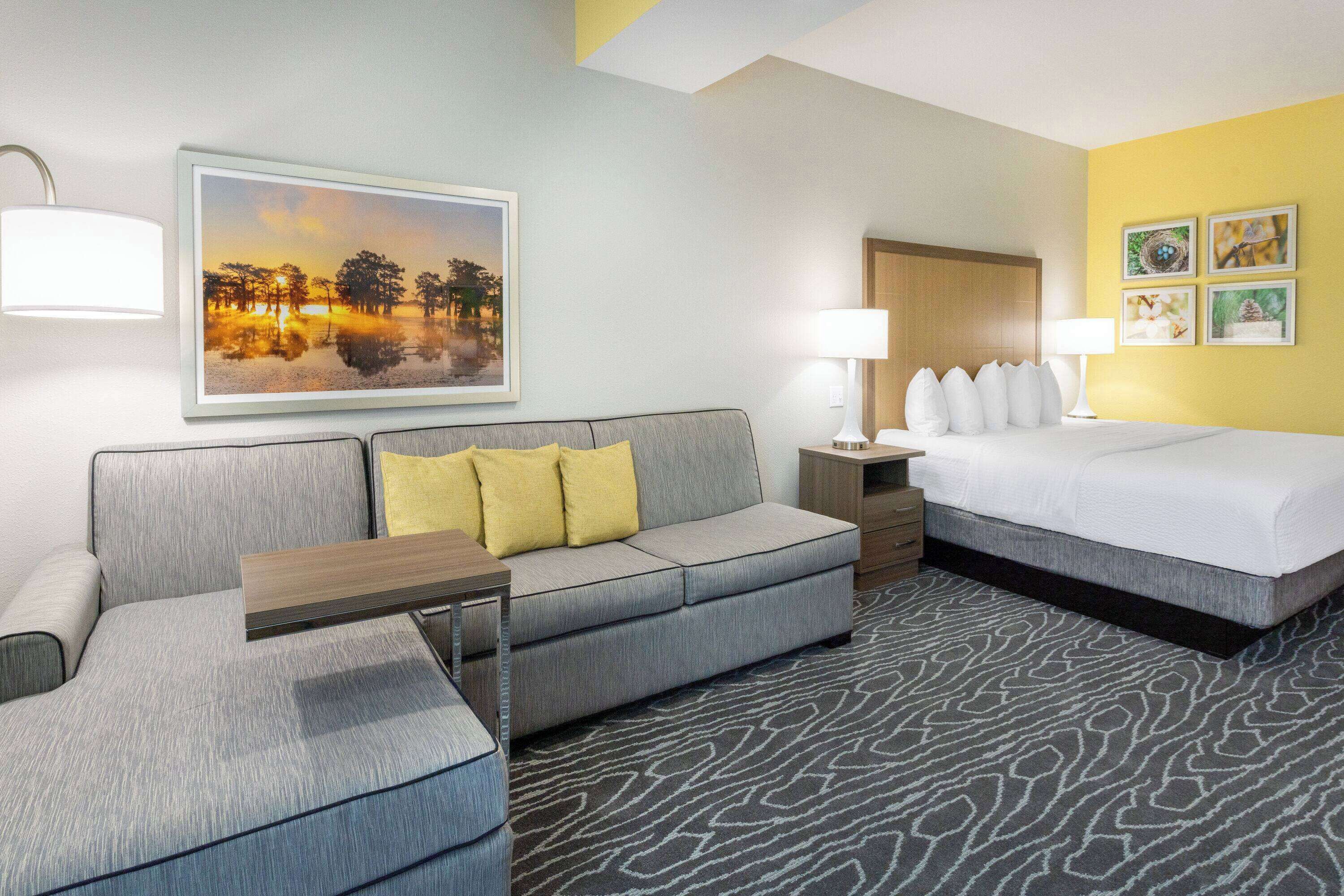 A hotel room with a grey sofa, a bed with white bedding, a wooden nightstand, and a wall-mounted lamp.