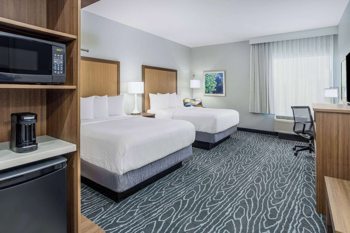 Een hotelkamer met twee bedden, een magnetron, een koffiezetapparaat, een bureau en een groot raam met gordijnen.