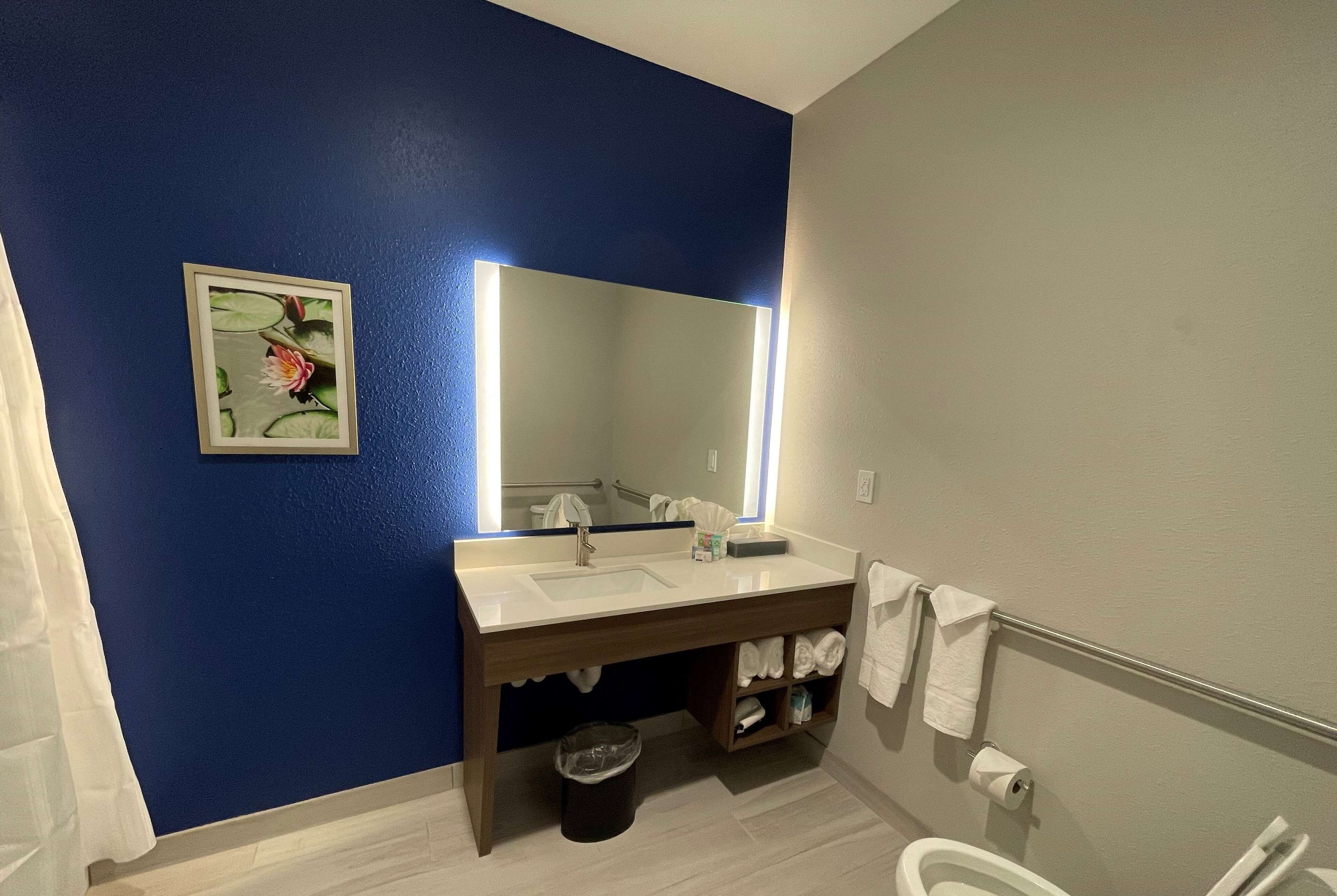 A bathroom with a blue wall, a white sink, a mirror with a light, a trash bin, and a toilet.