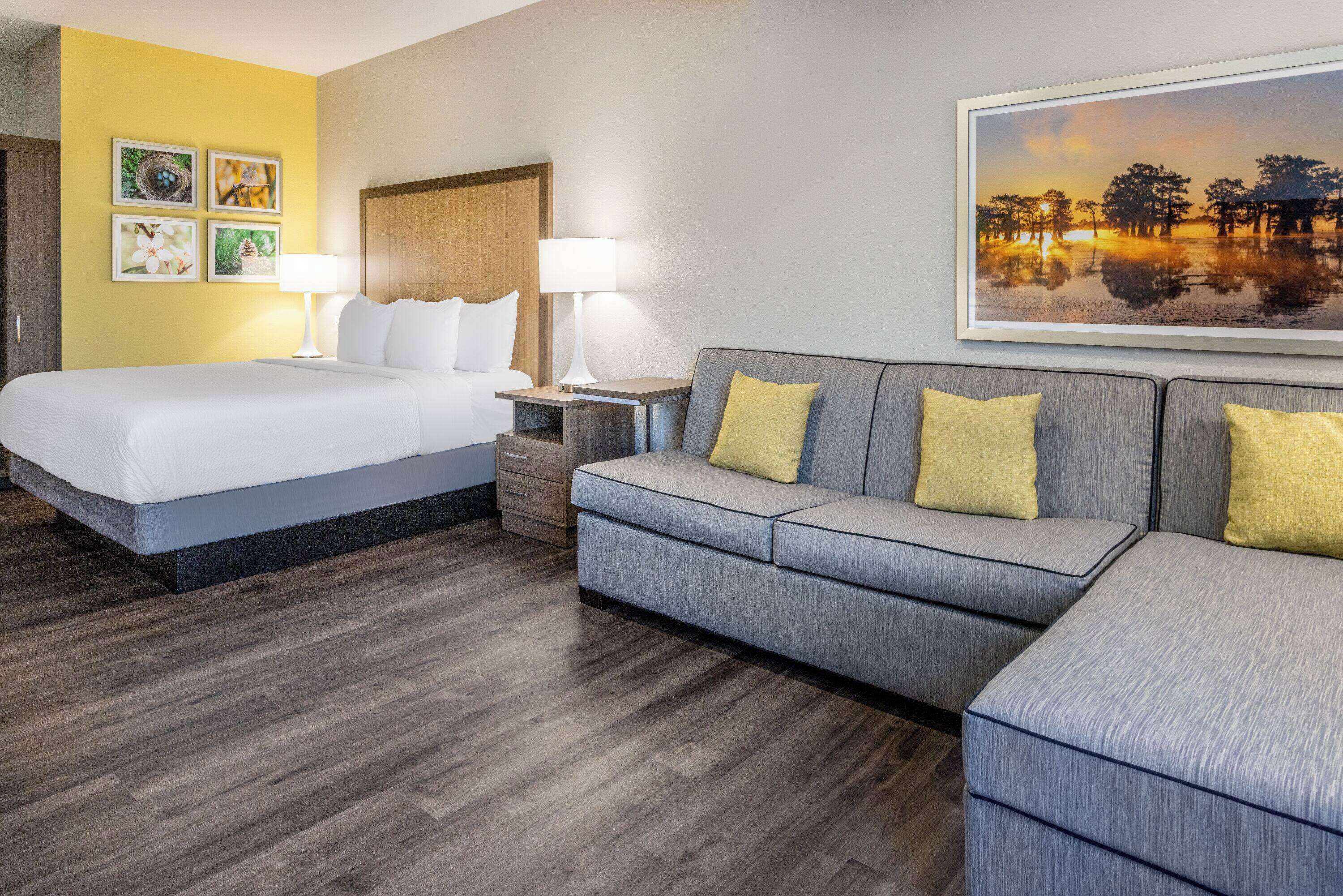 A hotel room with a bed, a gray sofa with yellow pillows, a nightstand with lamps, and a painting of a sunset over water on the wall.