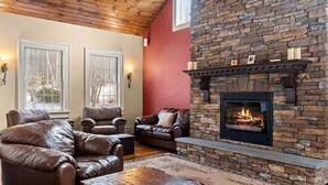 A cozy living room with leather sofas, a stone fireplace, and two windows with white frames. - Luxury 5-BR Mountain Estate 5-Min to Okemo (Ludlow)