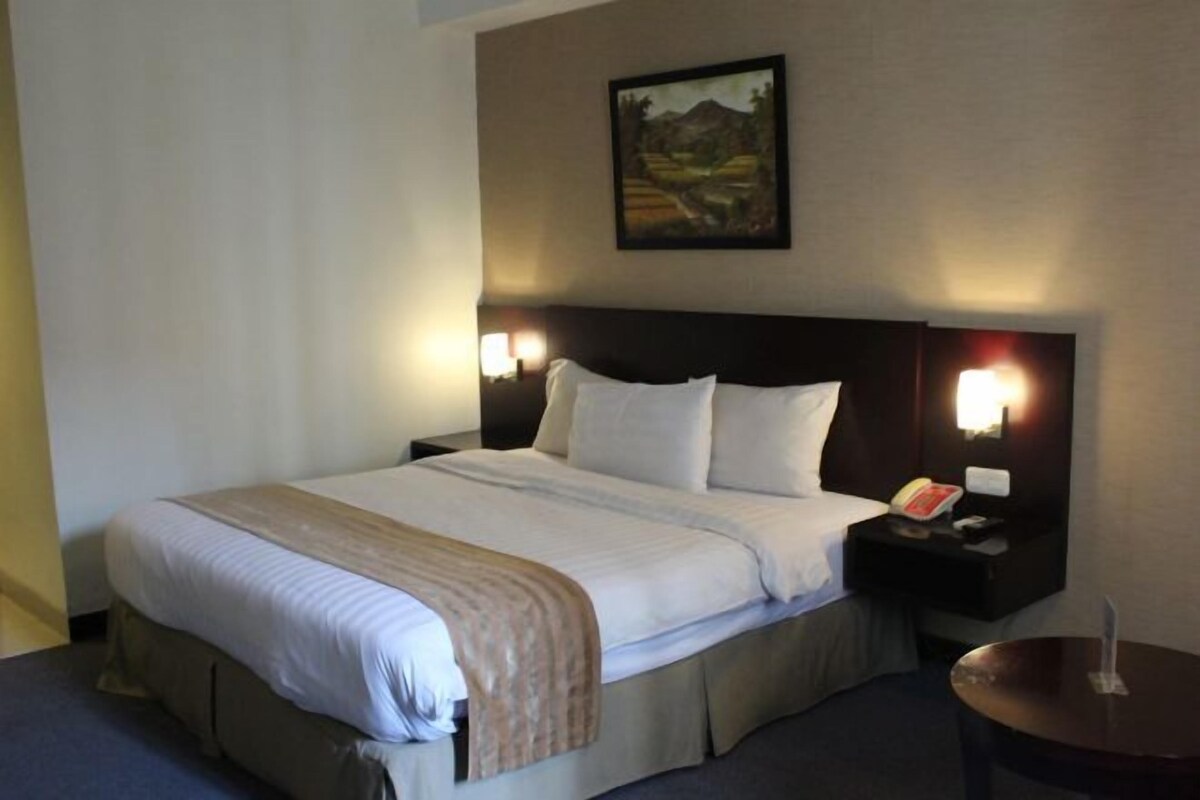 Habitación de hotel con una cama grande, dos mesitas de noche con lámparas, un cuadro en la pared y una pequeña mesa con un florero.