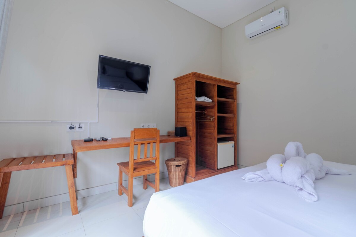 A hotel room with a bed, a wooden desk, a chair, a television mounted on the wall, a wardrobe, and a trash bin.