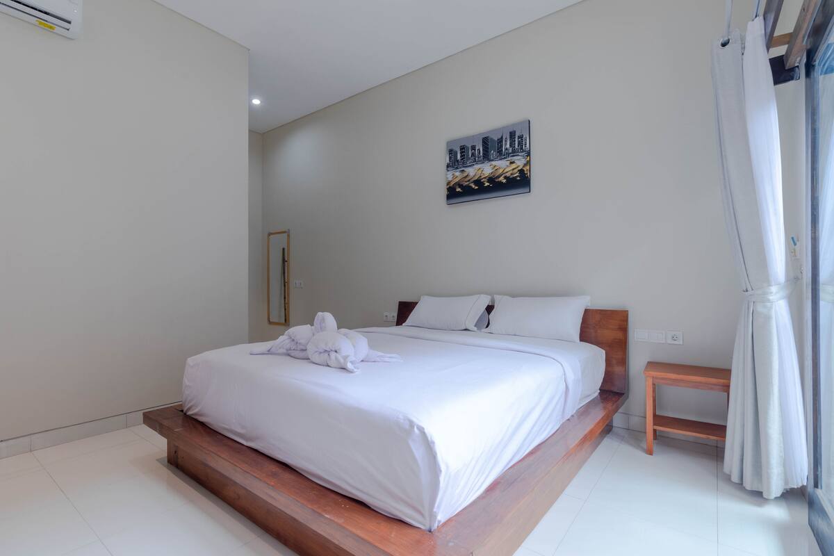 Een moderne slaapkamer met een groot bed, een houten bankje, een klein tafeltje en een stadsgezichtschilderij aan de muur.
