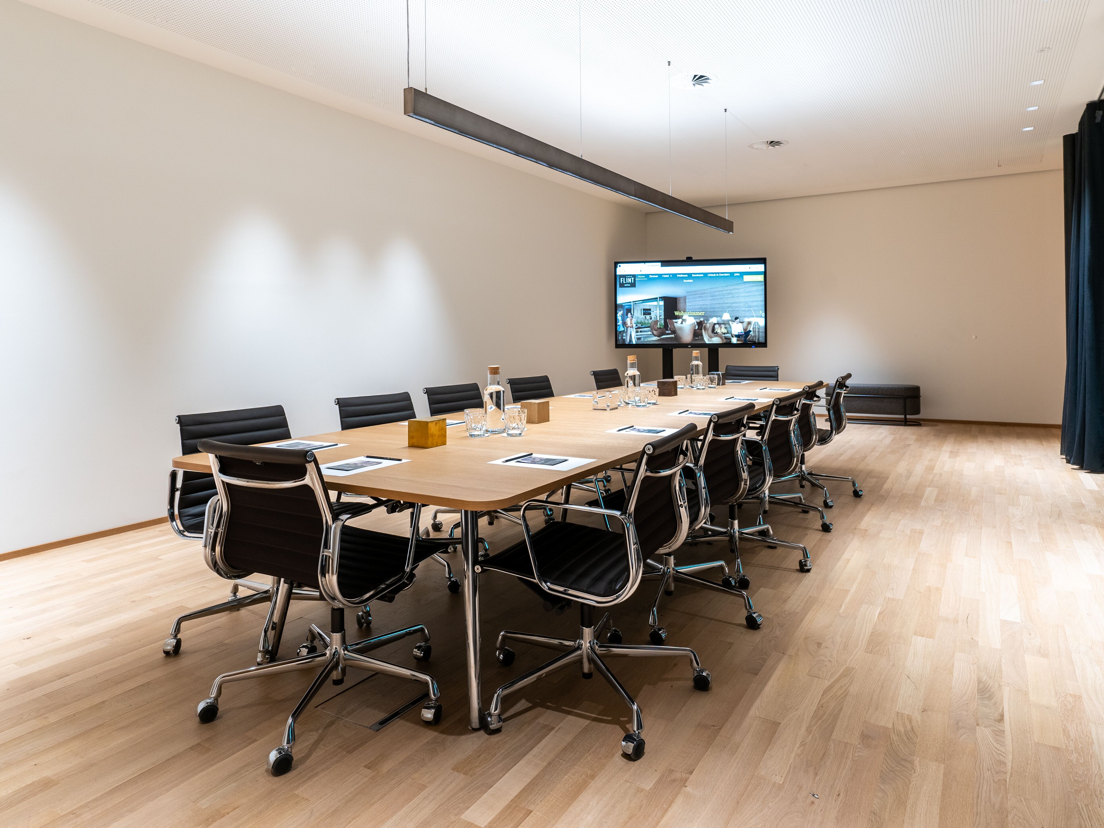 Een vergaderzaal met een lange tafel, zwarte stoelen, een groot scherm en een houten vloer.