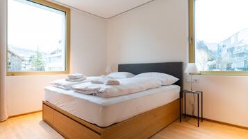 Ein modernes Schlafzimmer mit einem hölzernen Bett, weißer Bettwäsche und Blick auf Gebäude durch große Fenster.