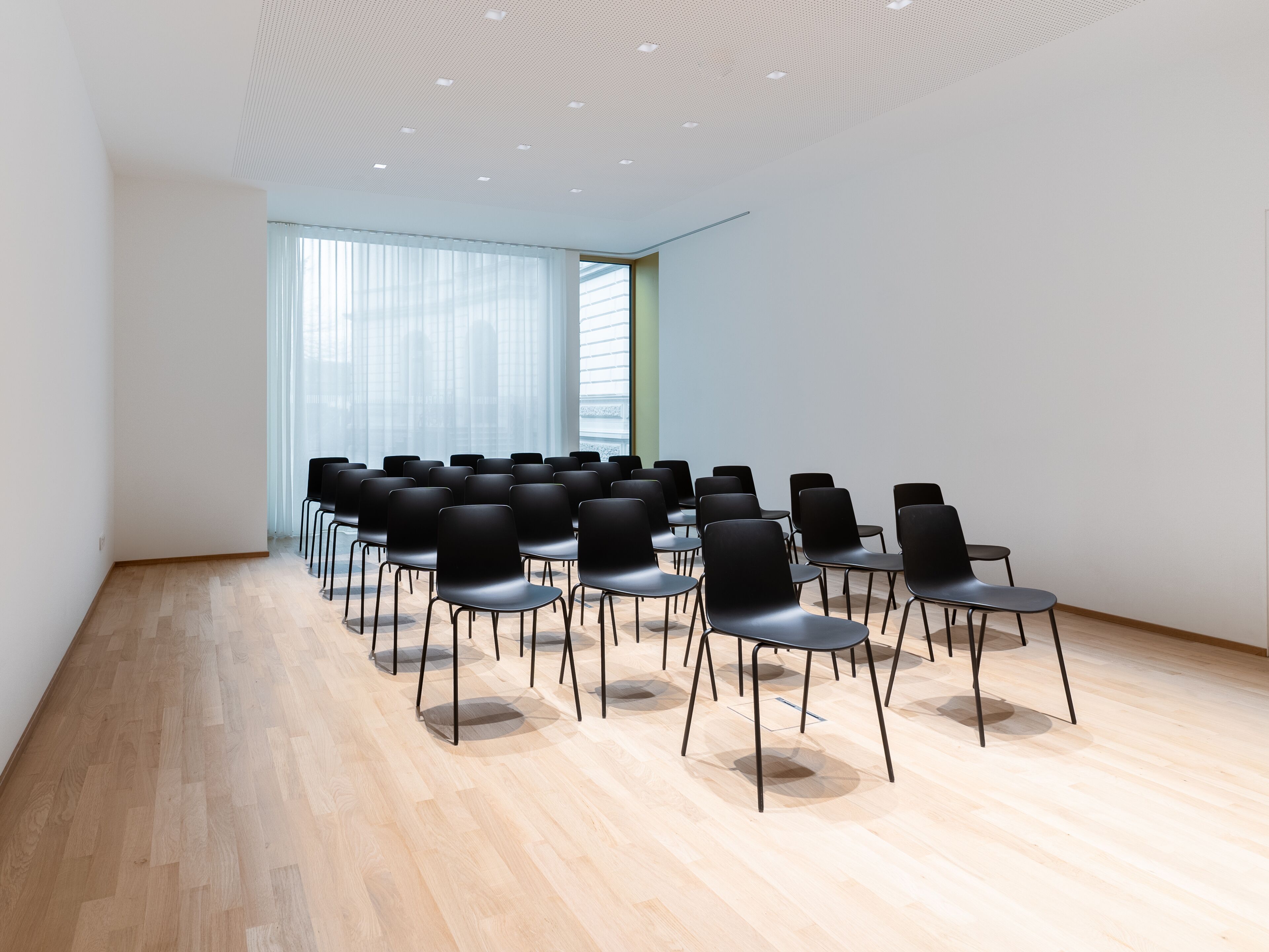 A conference room with rows of black chairs, wooden flooring, and a white ceiling with recessed lighting.
