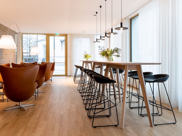 A modern dining area with a long wooden table, black bar stools, and orange chairs. - Hotel Flint  (Dornbirn)