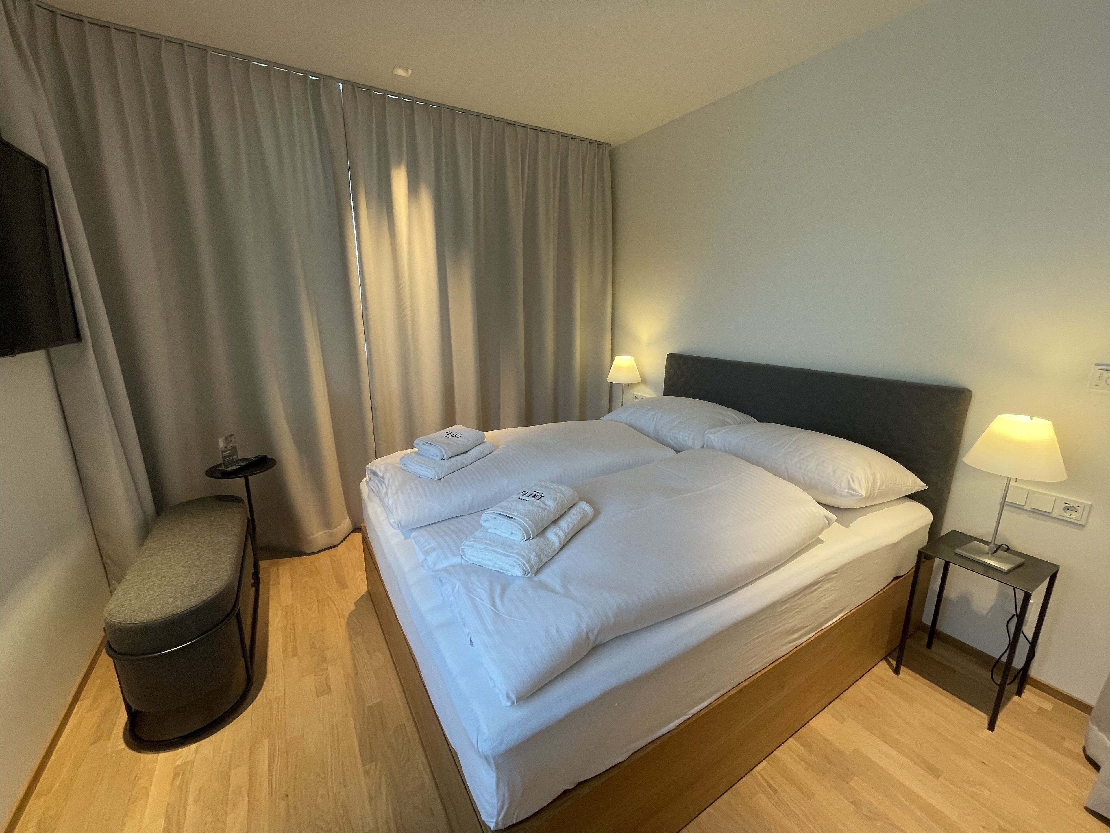 A hotel room with a large bed, two bedside tables with lamps, a small round table, and a bench.