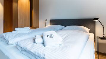 Cama bem arrumada com lençóis brancos e uma toalha com a inscrição "FLINT HOTEL", um guarda-roupa de madeira, uma mesa de cabeceira com abajur e cabeceira cinza.