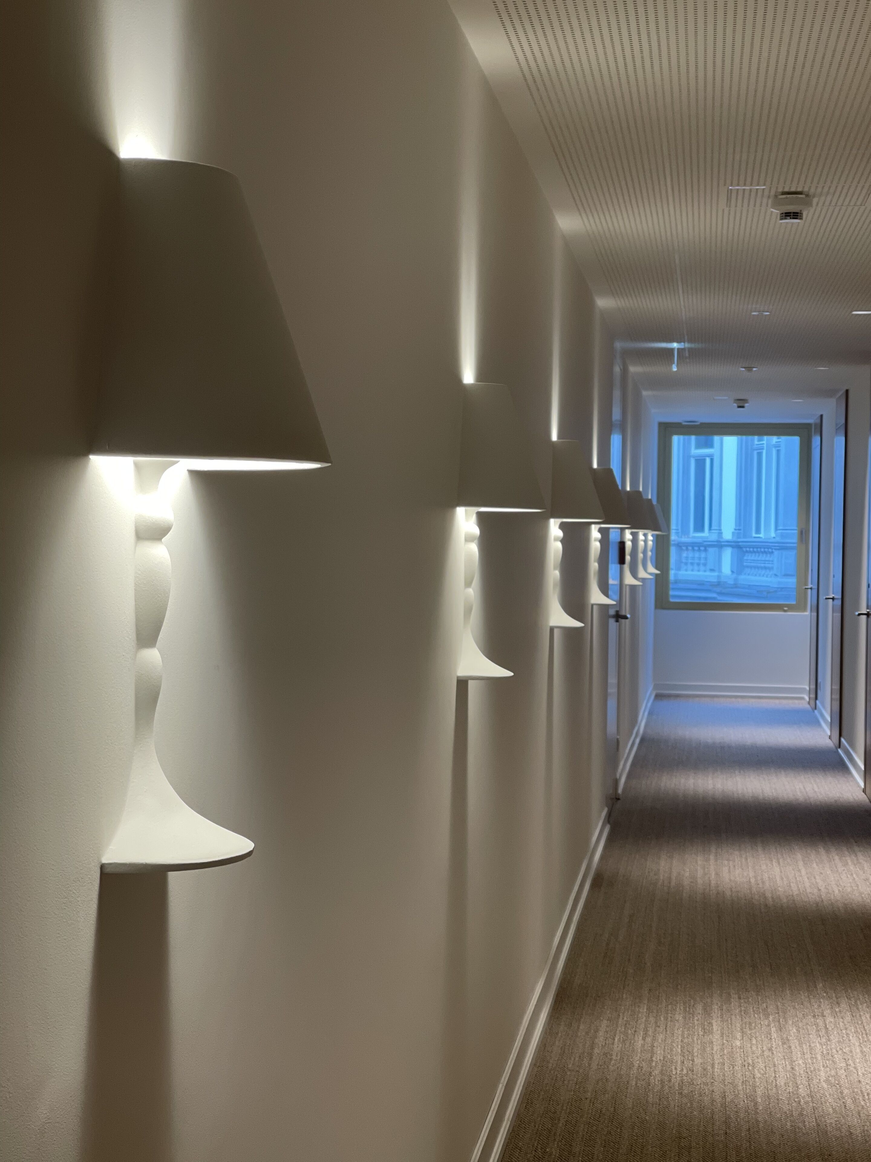 Un pasillo con lámparas modernas empotradas en la pared y piso alfombrado, que conduce a una puerta con vista a un edificio.