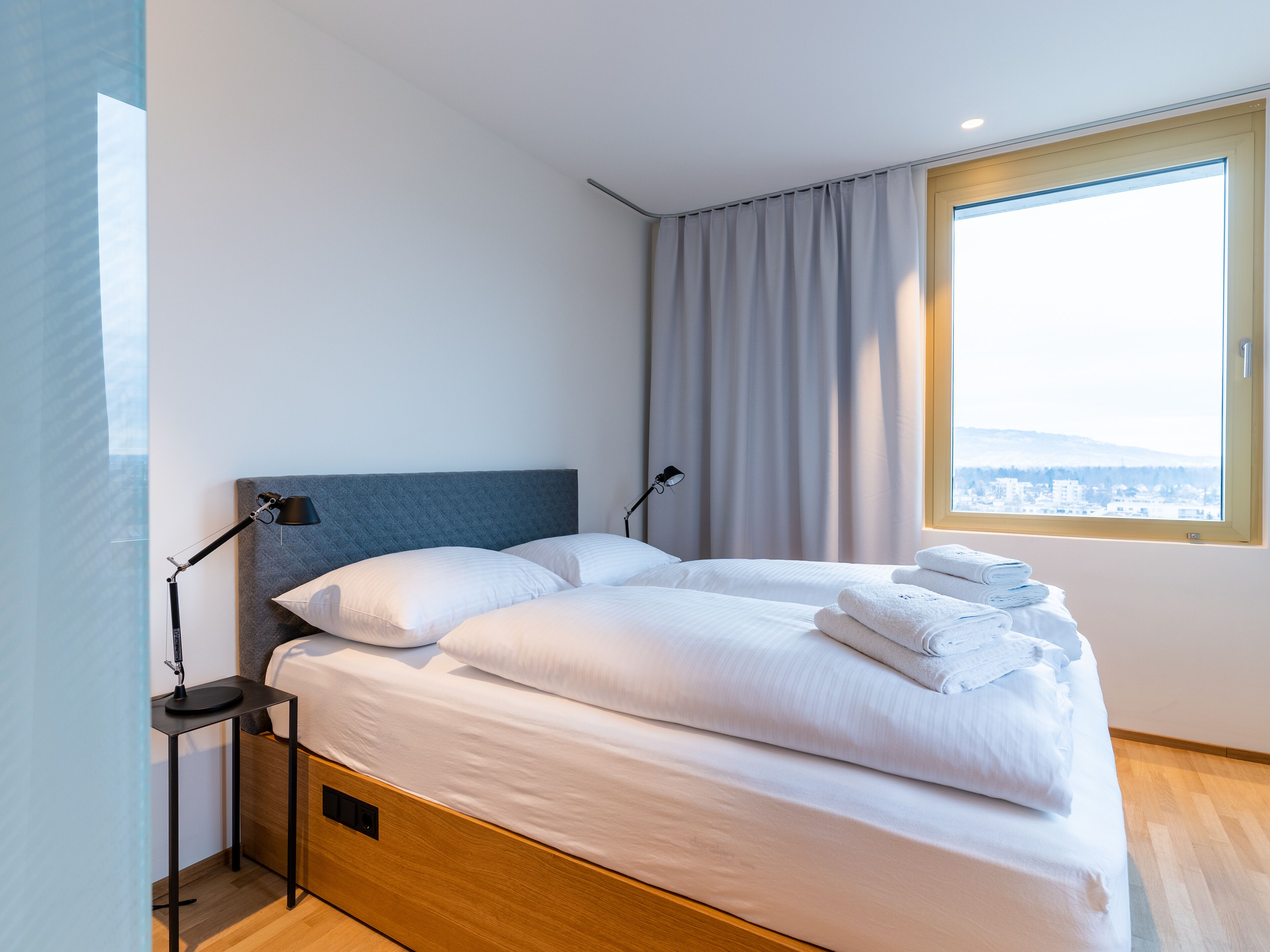 A modern hotel room with a large bed, a bedside table with a lamp, and a window with a city view.