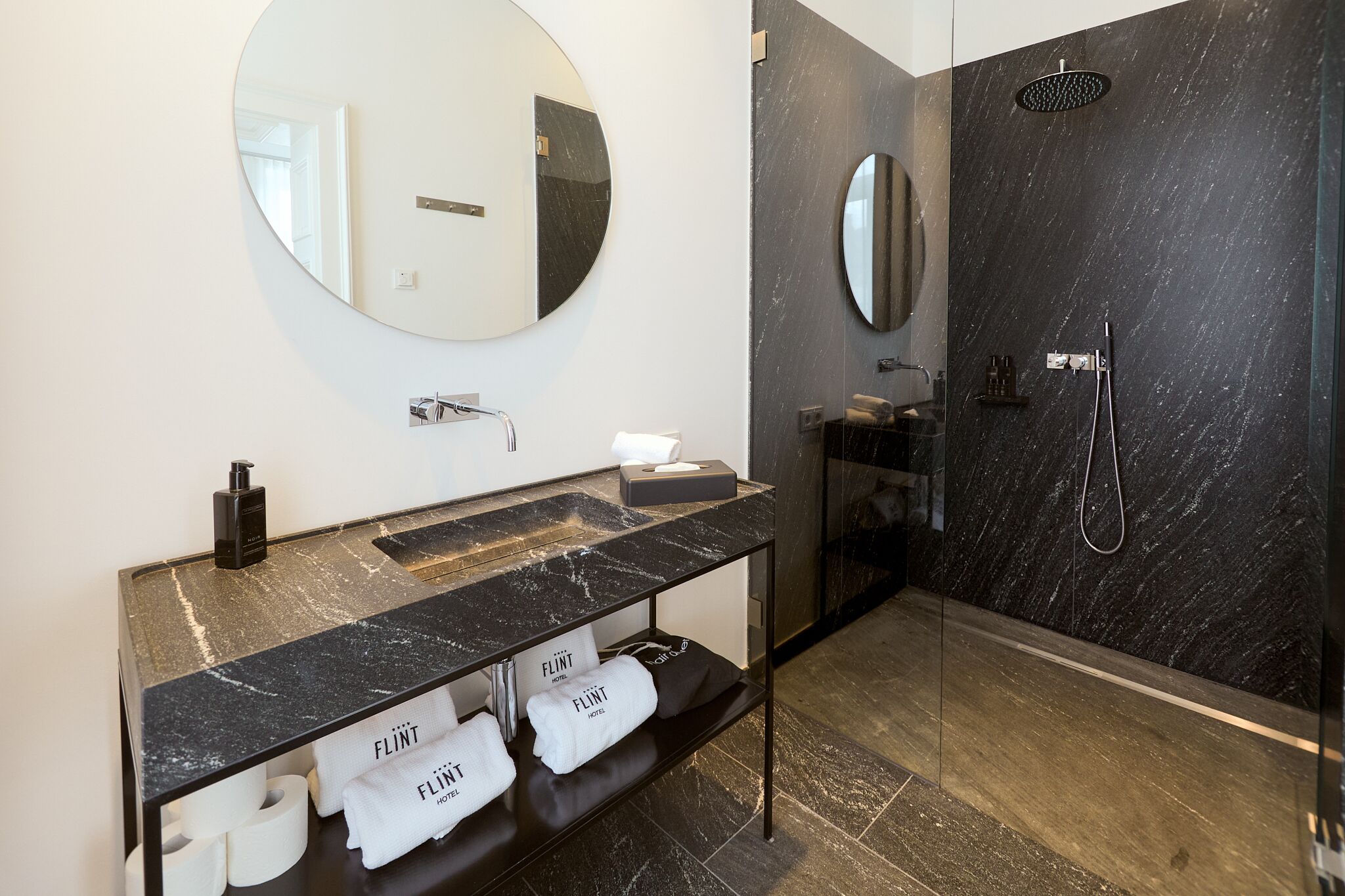 A modern bathroom with a marble vanity, a round mirror, a glass-enclosed shower, and towels labeled "FLINT HOTEL."